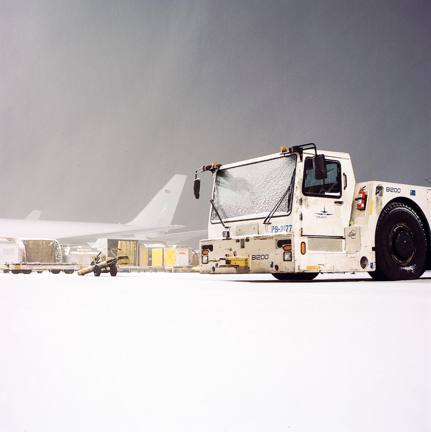 A snow-covered tow vehicle is parked on an airfield with an airplane and cargo containers in the background.