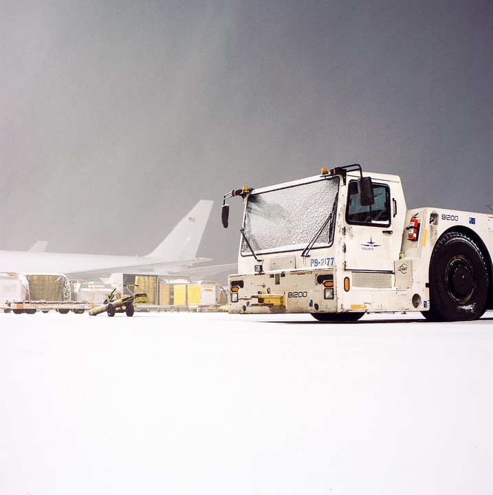 A snow-covered tow vehicle is parked on an airfield with an airplane and cargo containers in the background.