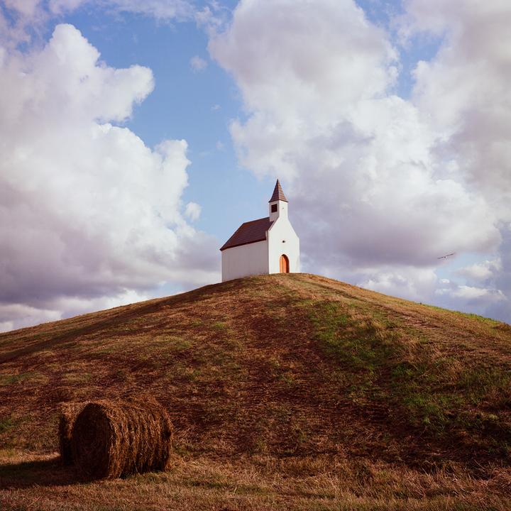 A small white chapel with a brown roof and steeple stands on top of a grassy hill under a cloudy sky, with a hay bale in the foreground.