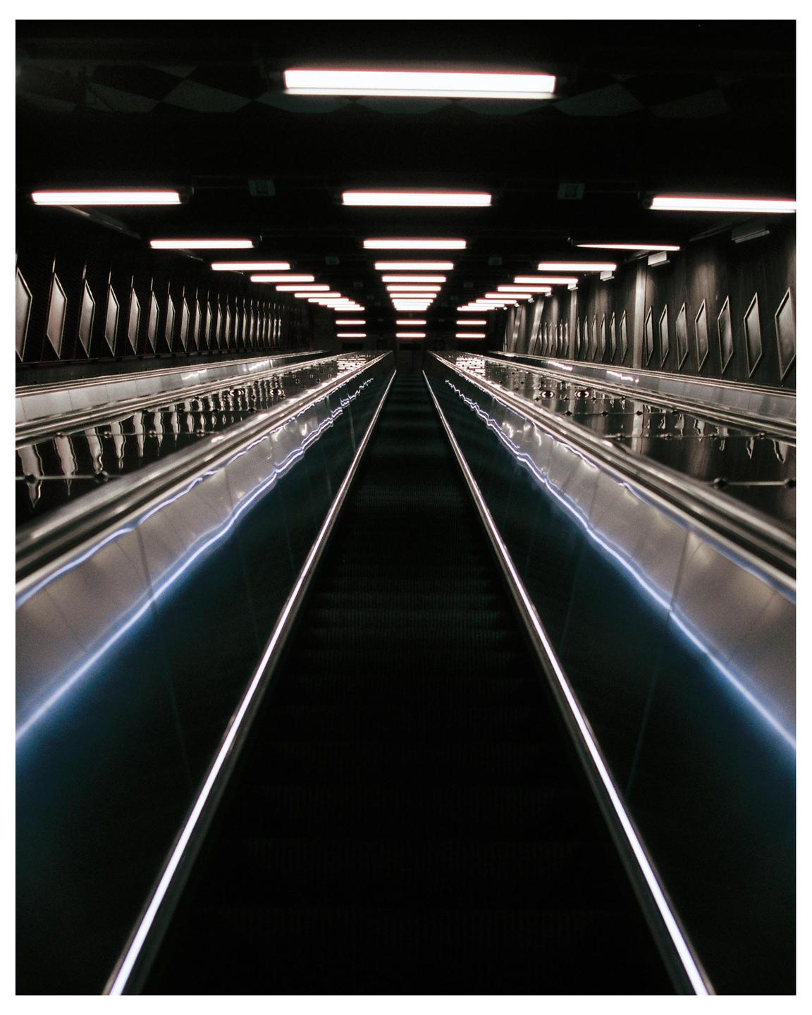 A long, dark escalator tunnel is illuminated by rows of fluorescent lights on the ceiling and glowing handrails.
