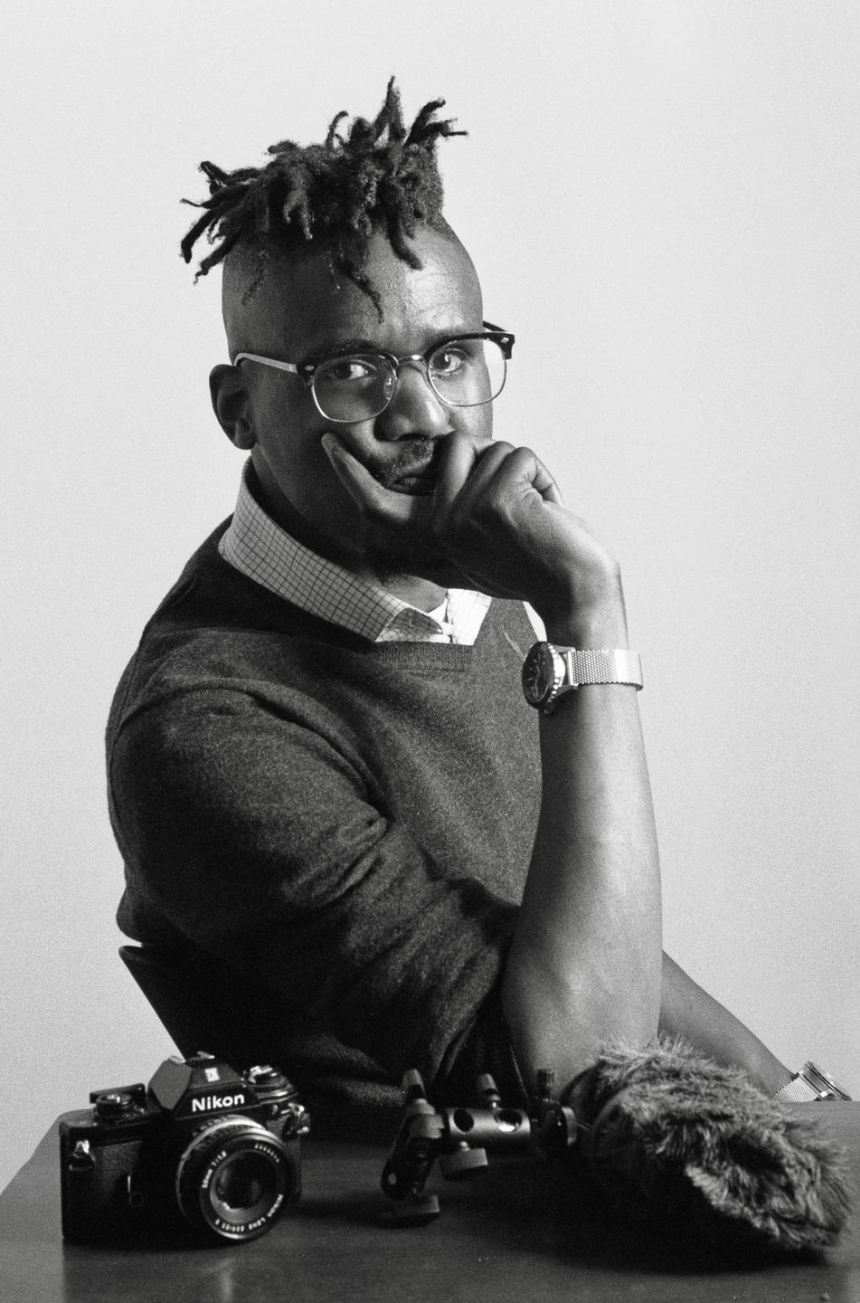A man with dreadlocks and glasses sits at a table with a camera and microphone.