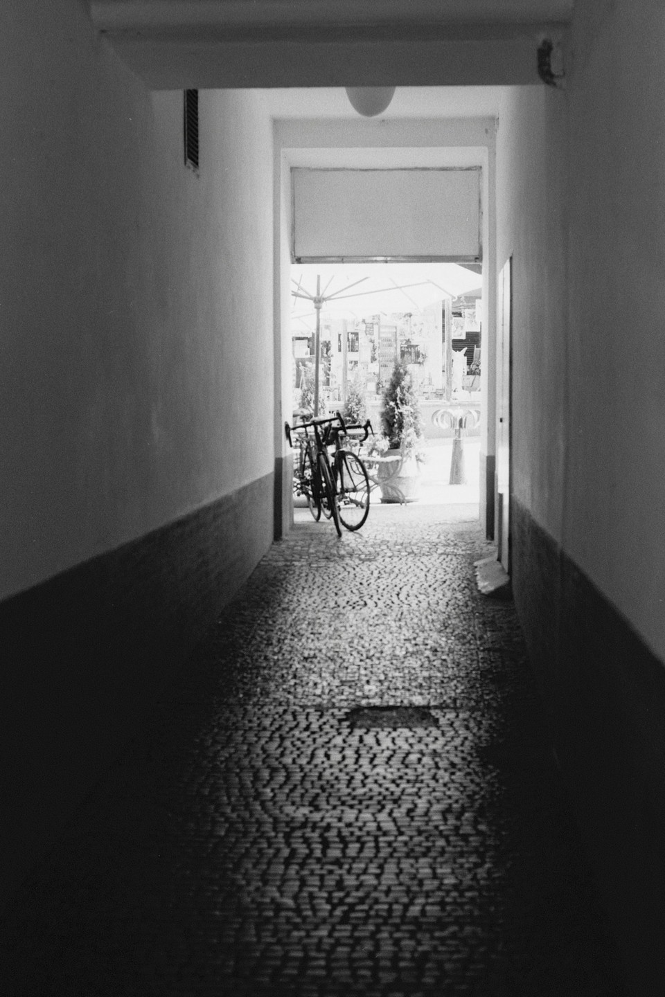 A bicycle is parked at the end of a narrow, cobblestone alleyway that opens to a bright outdoor area with an umbrella.