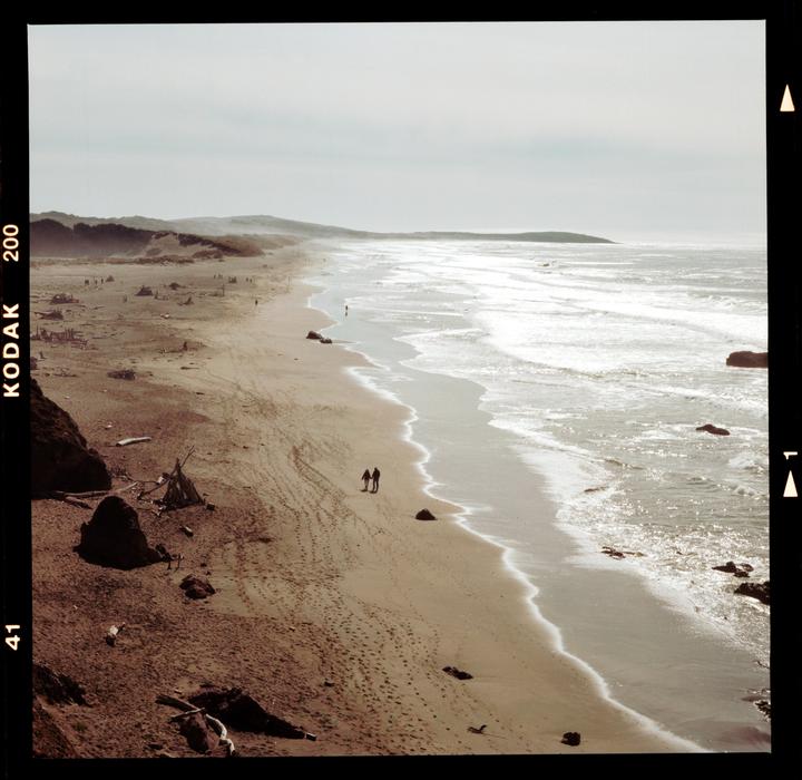 Two people walk along a wide sandy beach with waves crashing on the shore and hills in the distance.