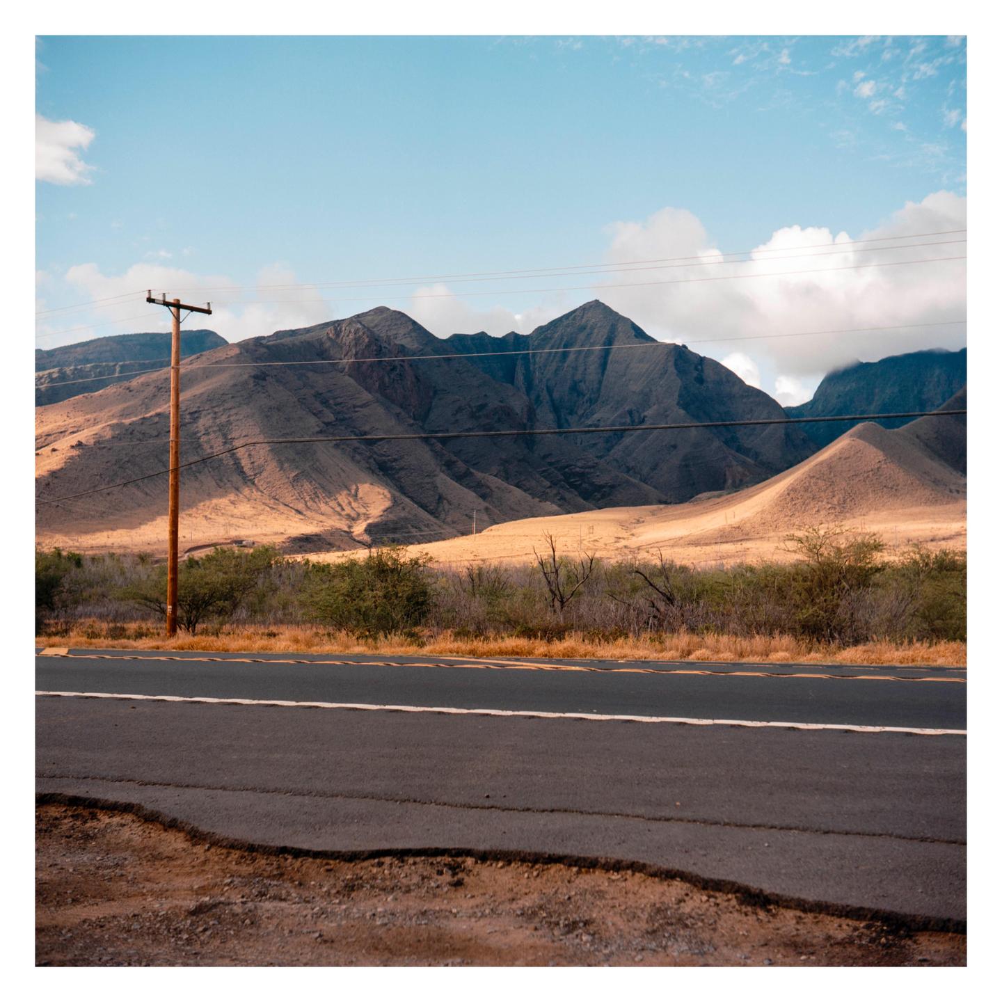 A road runs along the base of large, dry hills under a partly cloudy sky.