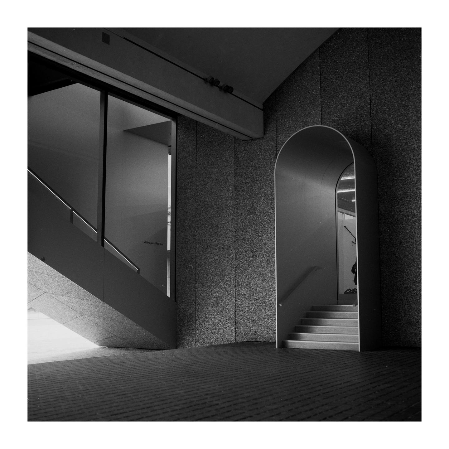 A black and white image shows a modern interior with a staircase on the left and an arched doorway with stairs on the right.
