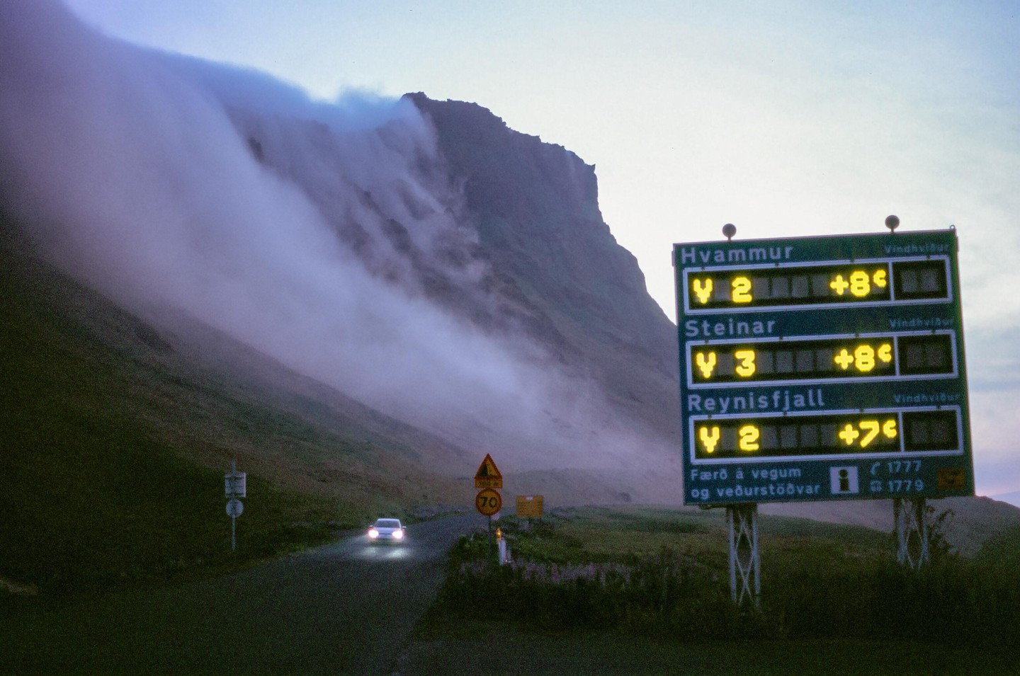 A car with its headlights on drives on a road next to a mountain covered in fog and a large sign displaying information.