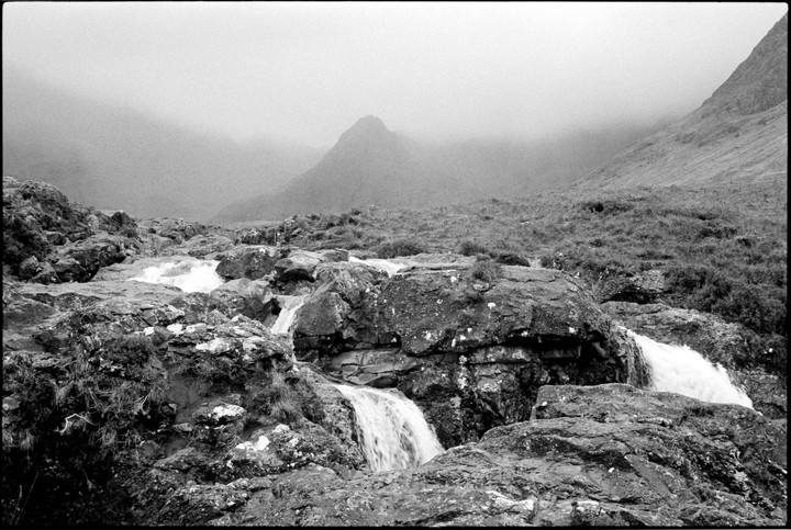 A stream cascades over rocks in a mountainous, hazy landscape.