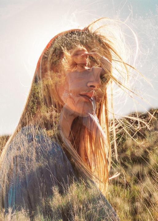 A double exposure shows a woman with long hair in a field of tall grass, with a second exposure of a woman shielding her eyes from the sun superimposed over her face.