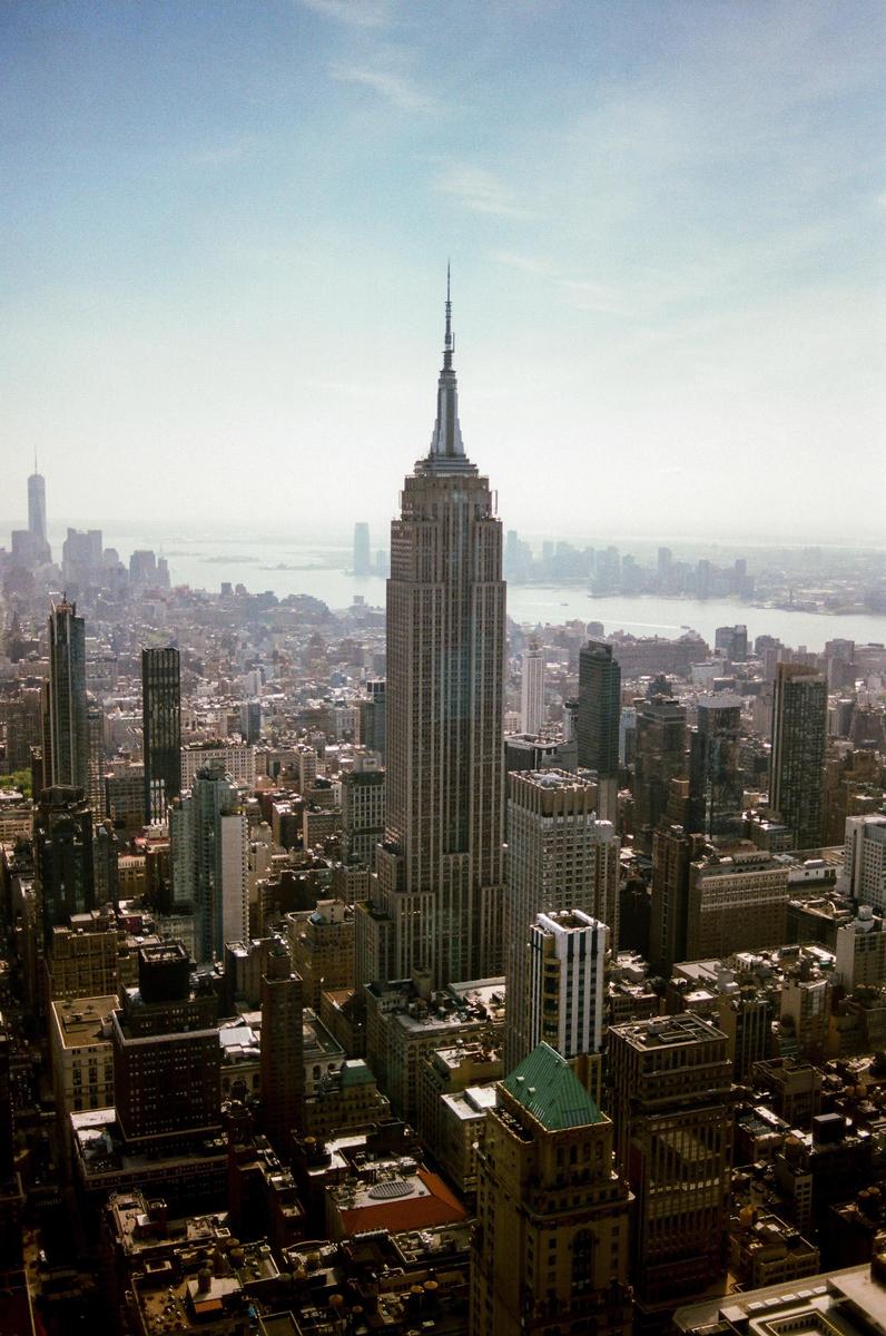 The Empire State Building rises above the surrounding cityscape with a body of water and distant land visible in the background.