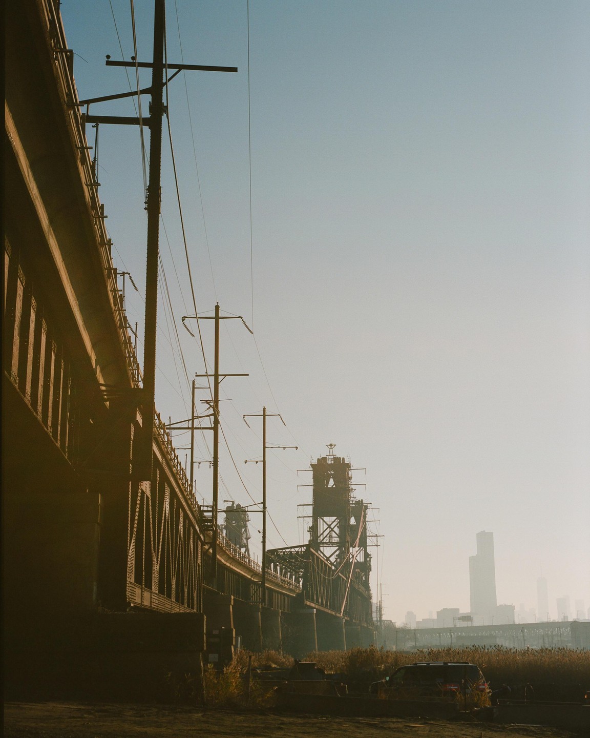 A long bridge with power lines runs alongside a hazy cityscape with tall buildings in the distance.