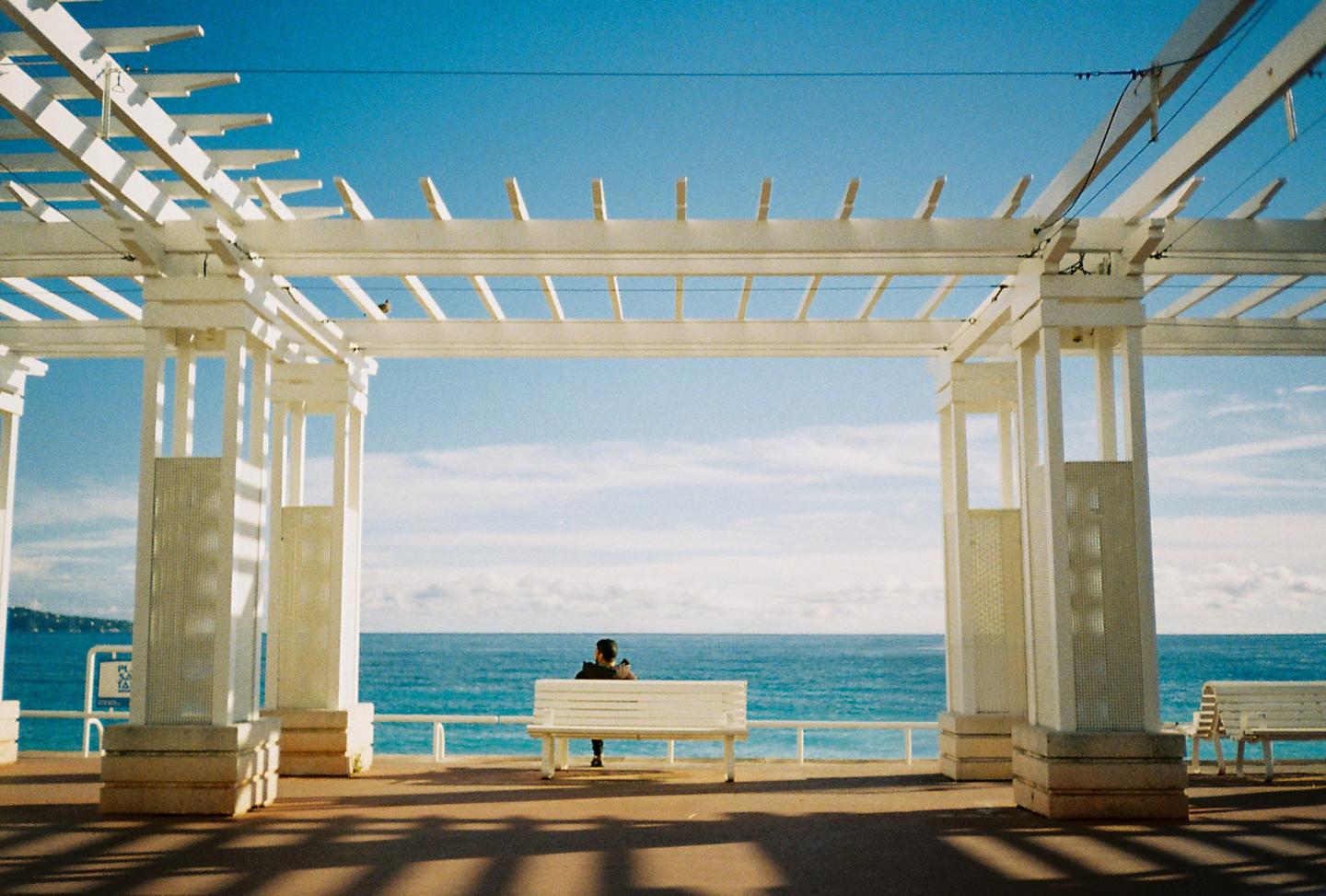 A person sits on a white bench under a white pergola, looking out at the blue sea.