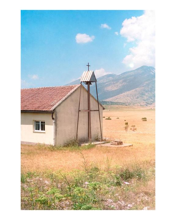 A small church with a bell tower stands in a dry, grassy field with mountains in the background under a blue sky.