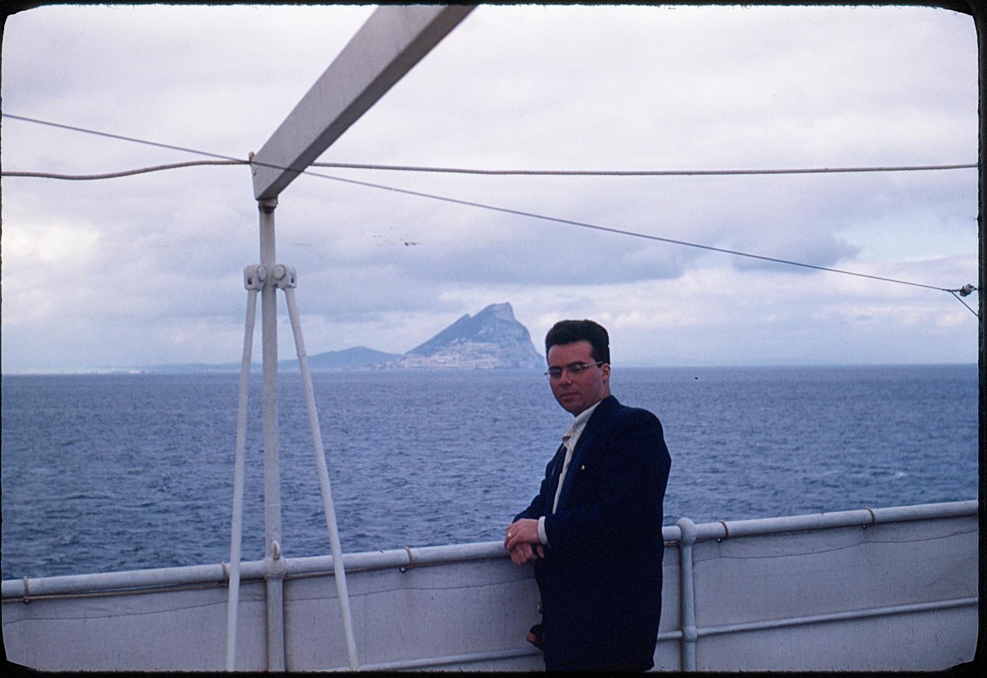 A man in a suit stands on a boat deck with the Rock of Gibraltar in the background.