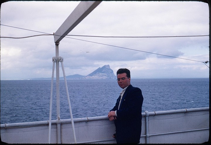A man in a suit stands on a boat deck with the Rock of Gibraltar in the background.