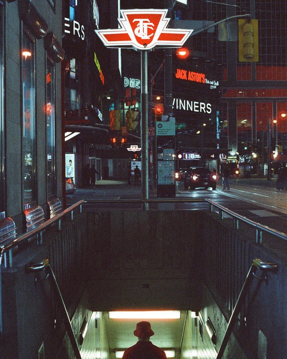 A person in a red hat descends into a subway station at night, with city lights and buildings visible above.