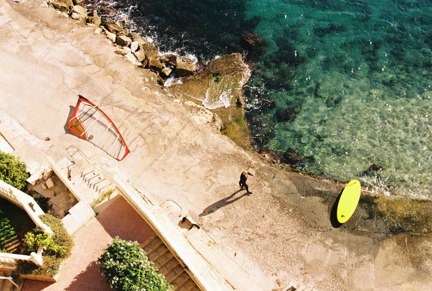 A person in a wetsuit walks along a concrete path next to the ocean, with a windsurf sail and a surfboard on the ground.