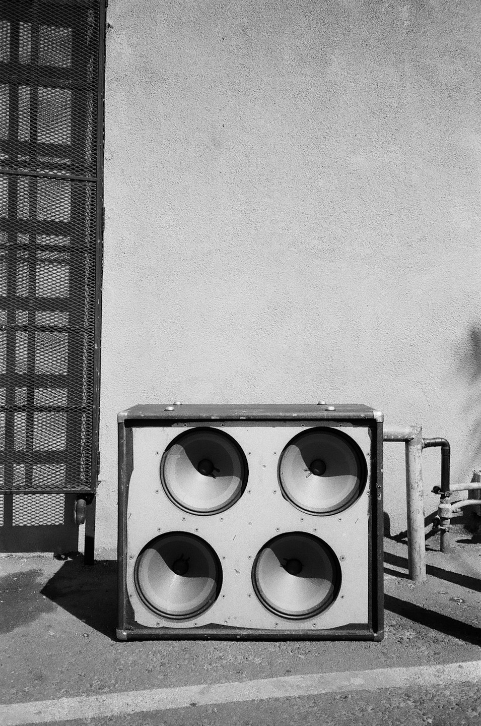 A large speaker cabinet with four speakers sits on the ground next to a wall and a metal grate.