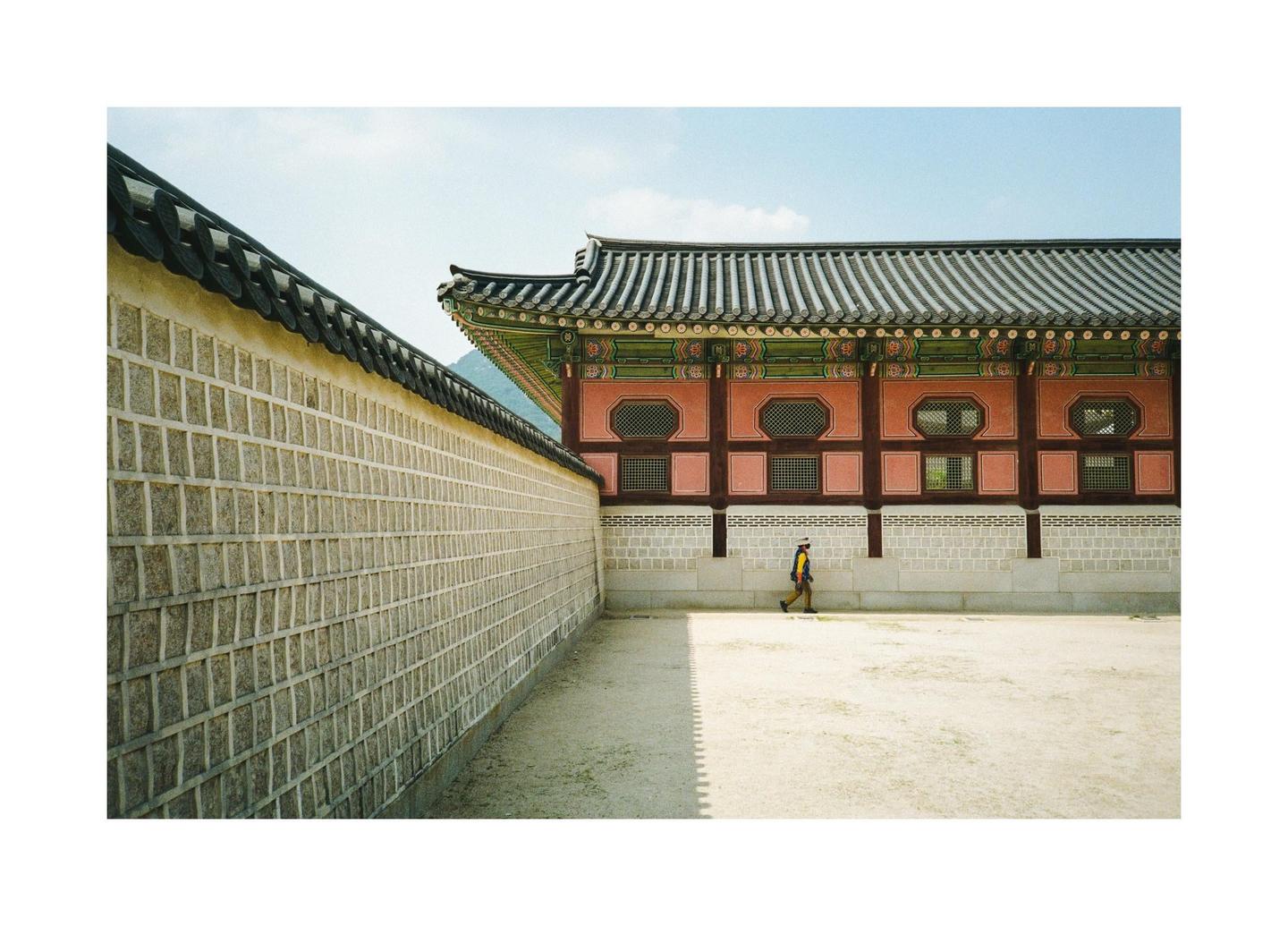 A person walks along a dirt path next to a stone wall and a traditional Korean building with a tiled roof.