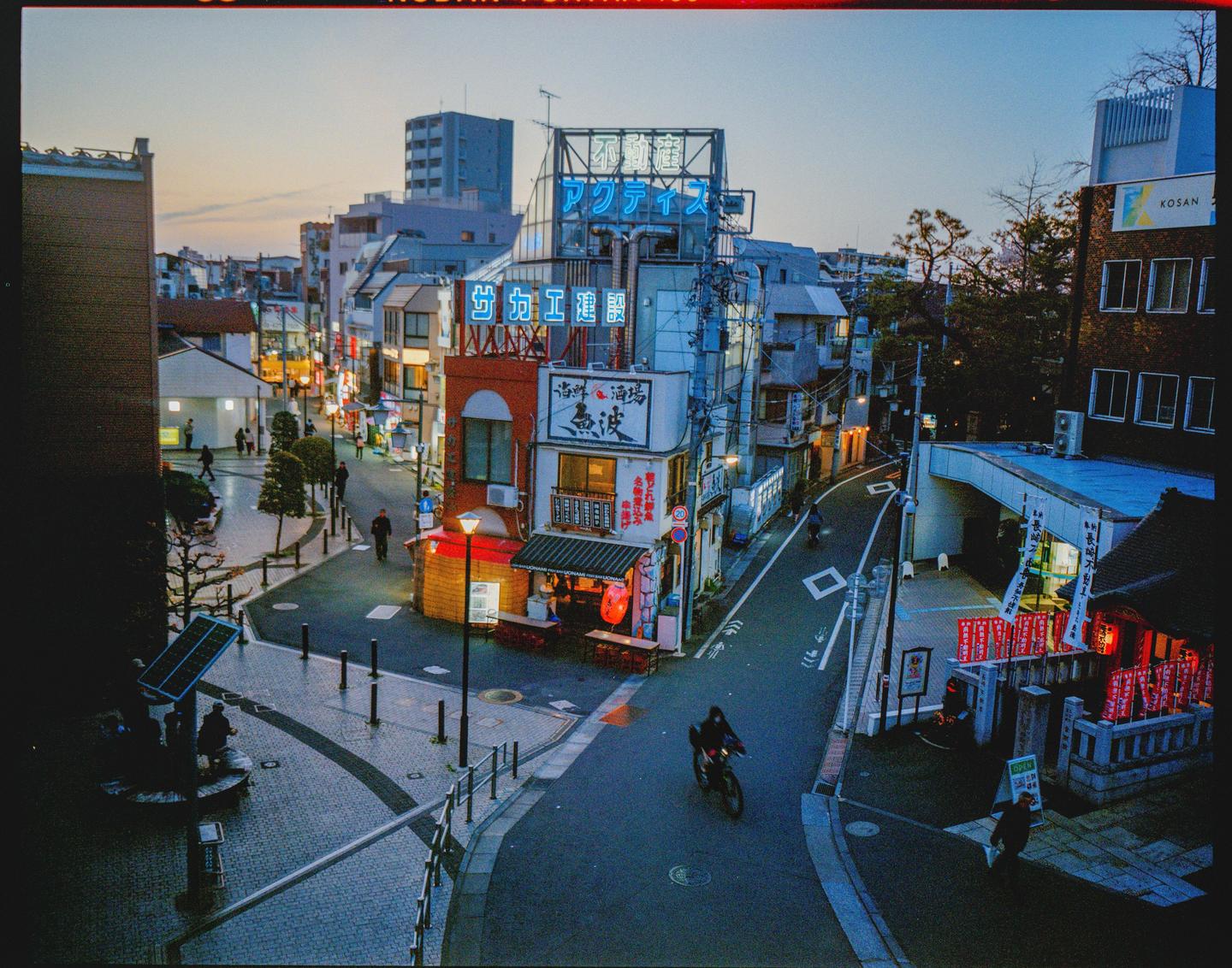 A street scene in a Japanese city at dusk shows buildings with neon signs and people walking and cycling.