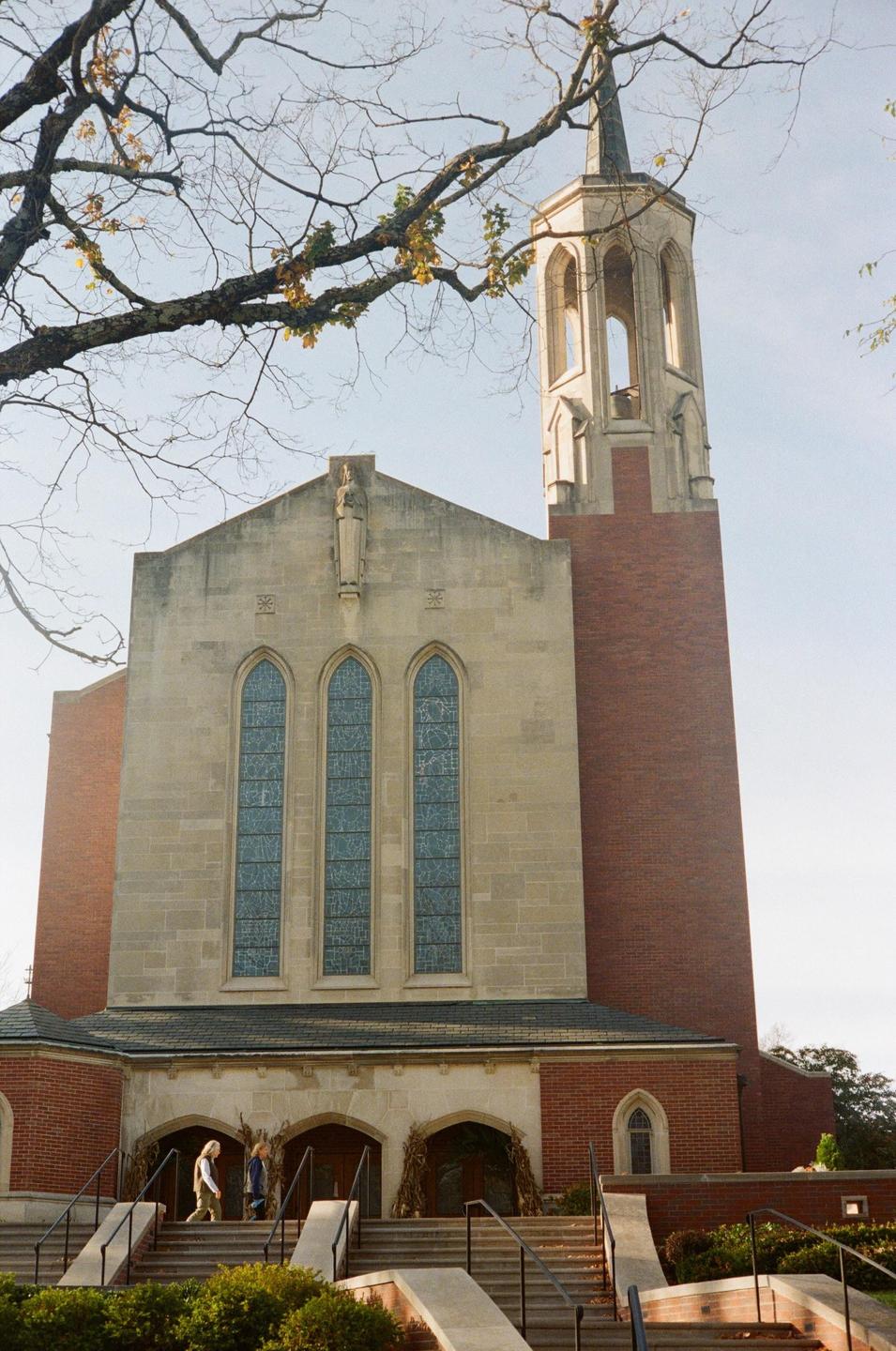A large brick and stone church with a tall bell tower stands under a clear sky, with bare tree branches in the foreground.
