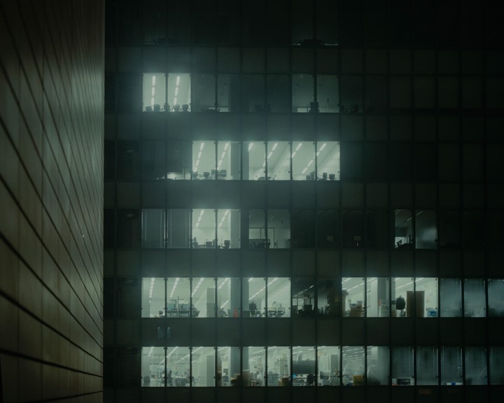A grid of illuminated office windows reveals various workspaces and a few people inside a tall building at night.