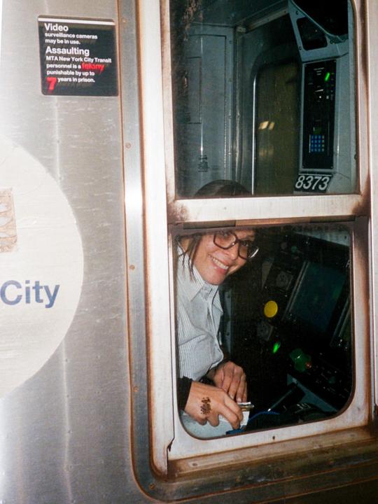 A woman wearing glasses and a striped shirt smiles from the window of a subway car, with a warning sign visible on the left.