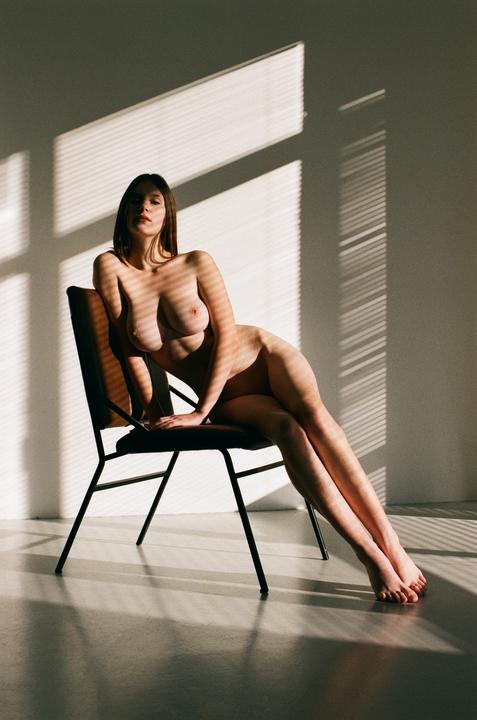 A nude woman sits on a chair with shadows from window blinds cast across her body and the wall behind her.