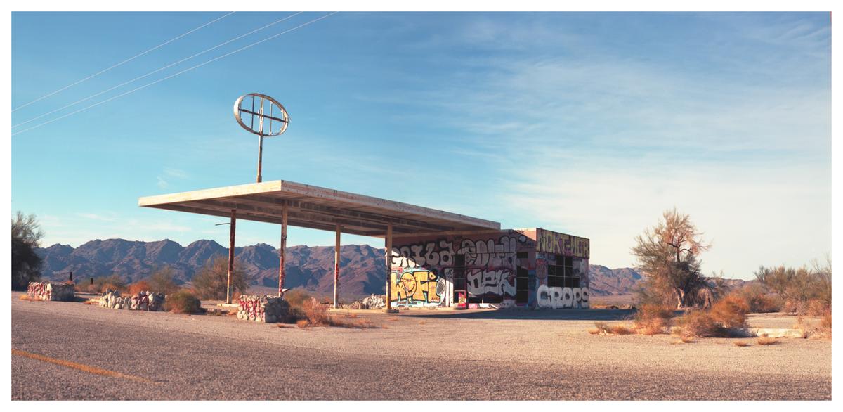 An abandoned gas station with graffiti on its walls stands in a desert landscape with mountains in the background.