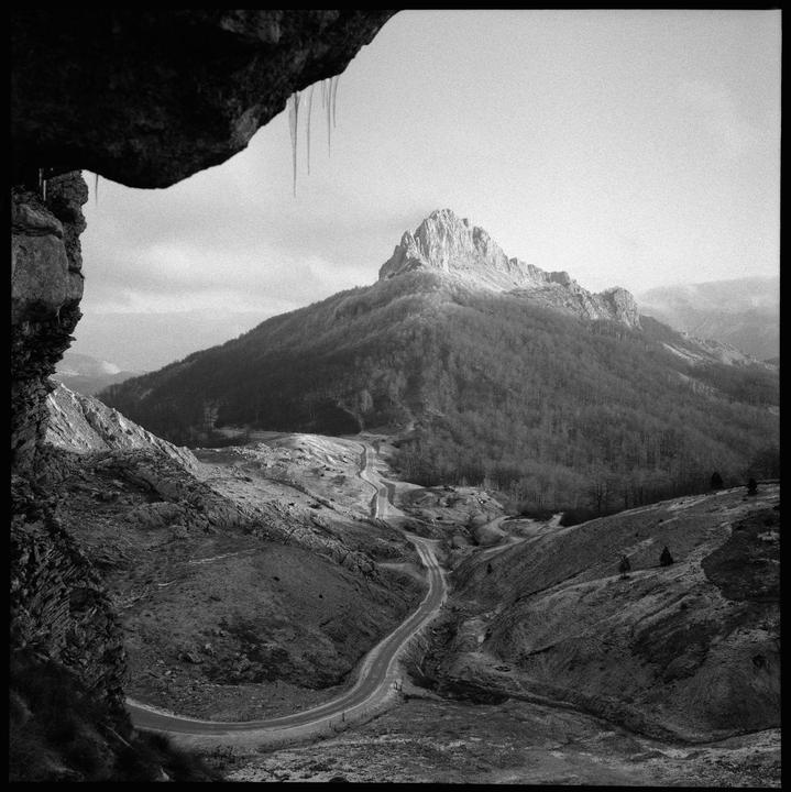 A winding road leads through a valley towards a large, rocky mountain peak under a cloudy sky, with icicles hanging from a rock formation in the foreground.