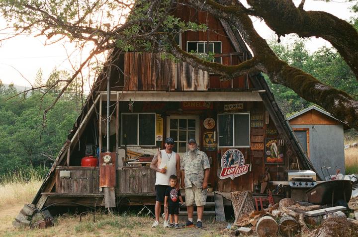 A man, a boy, and another man stand in front of a rustic wooden cabin with a large tree branch overhead.