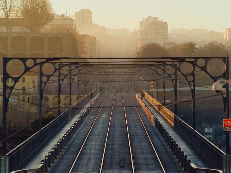 A bridge with train tracks and a pedestrian walkway extends into the distance under a hazy sky, with buildings visible on either side.
