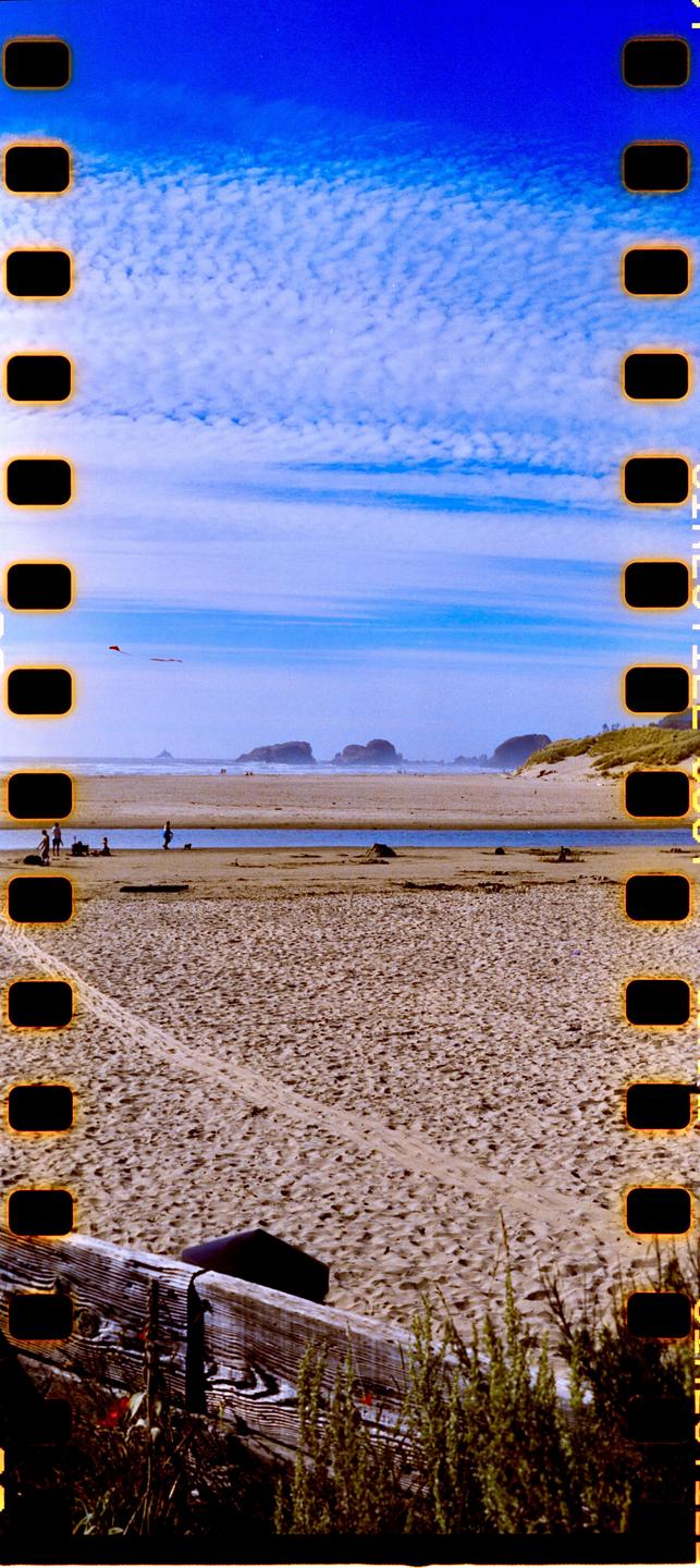 A panoramic view of a sandy beach with a stream, distant rock formations, and a bright blue sky with clouds.