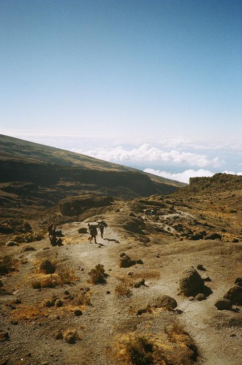 A group of people hike along a rocky, arid mountain path with clouds visible in the distance.