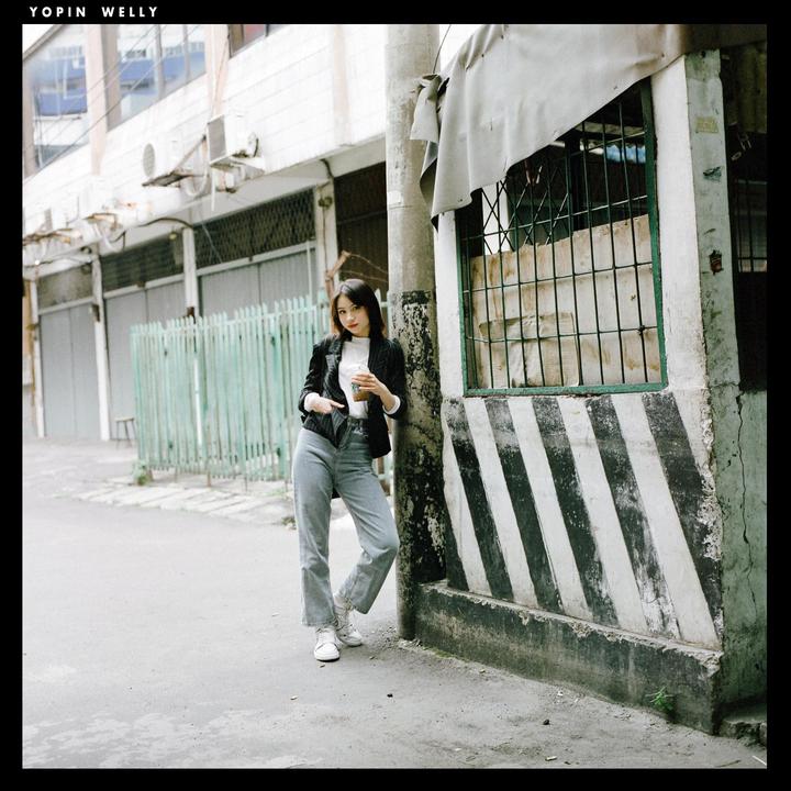 A woman stands on a street holding a drink, leaning against a concrete pillar with black and white stripes.