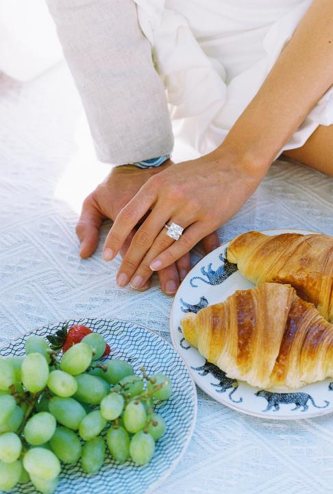 A couple's hands are clasped together on a white blanket next to plates of croissants and grapes.