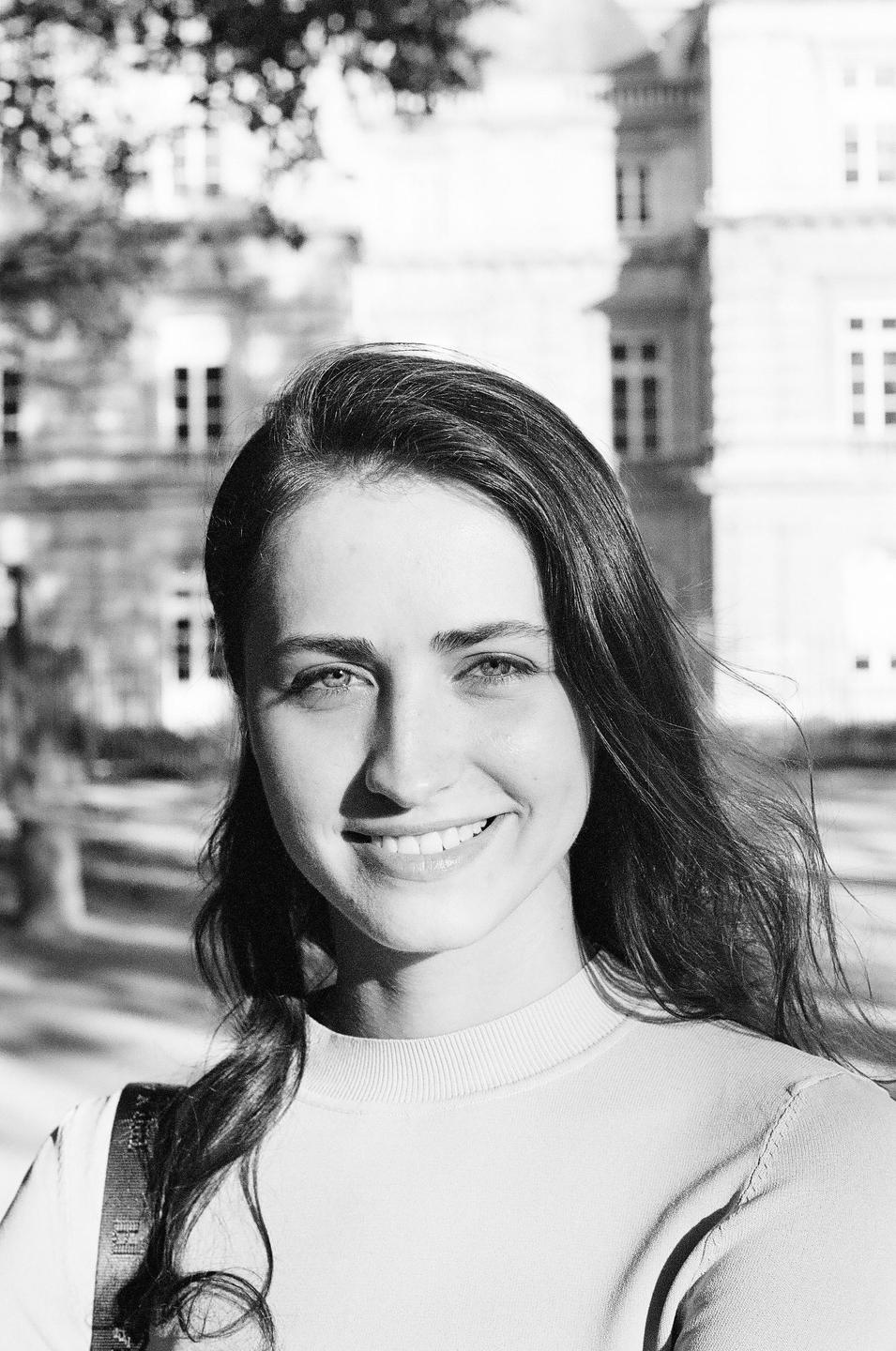 A smiling woman with long hair is shown in a close-up portrait with a building in the background.