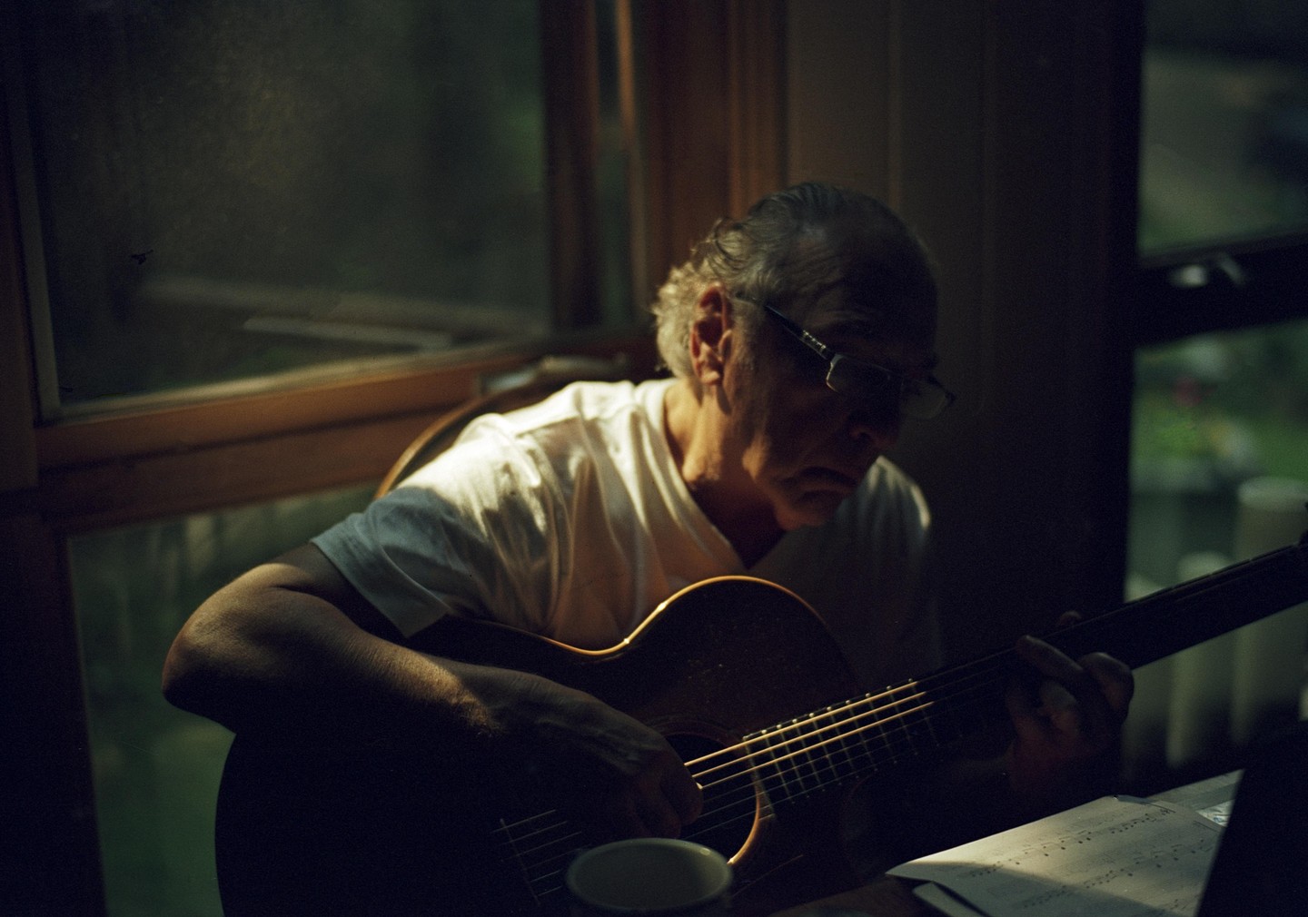 A man with glasses plays an acoustic guitar by a window, with sheet music visible on a table.