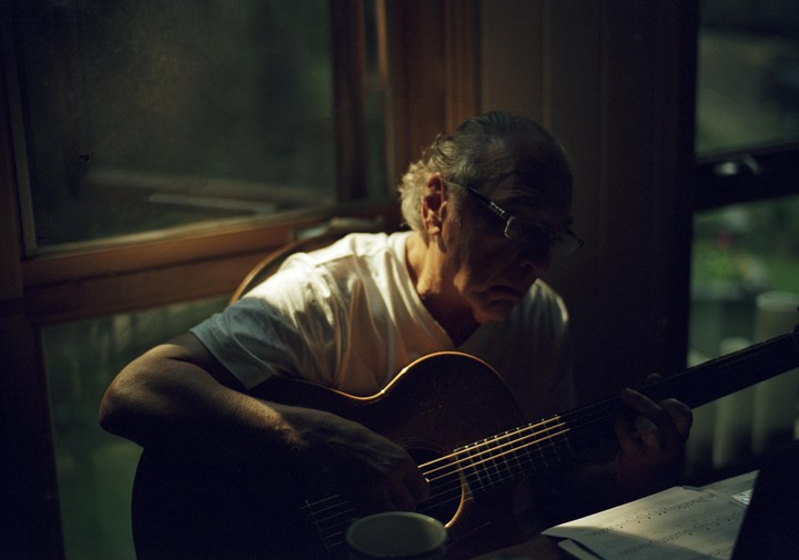 A man with glasses plays an acoustic guitar by a window, with sheet music visible on a table.