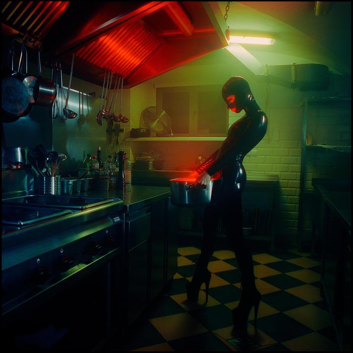 A person in a shiny black full-body suit and high heels holds a pot in a kitchen with red and green lighting.