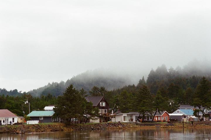 A row of houses sits along a body of water with a forested, foggy hill in the background.