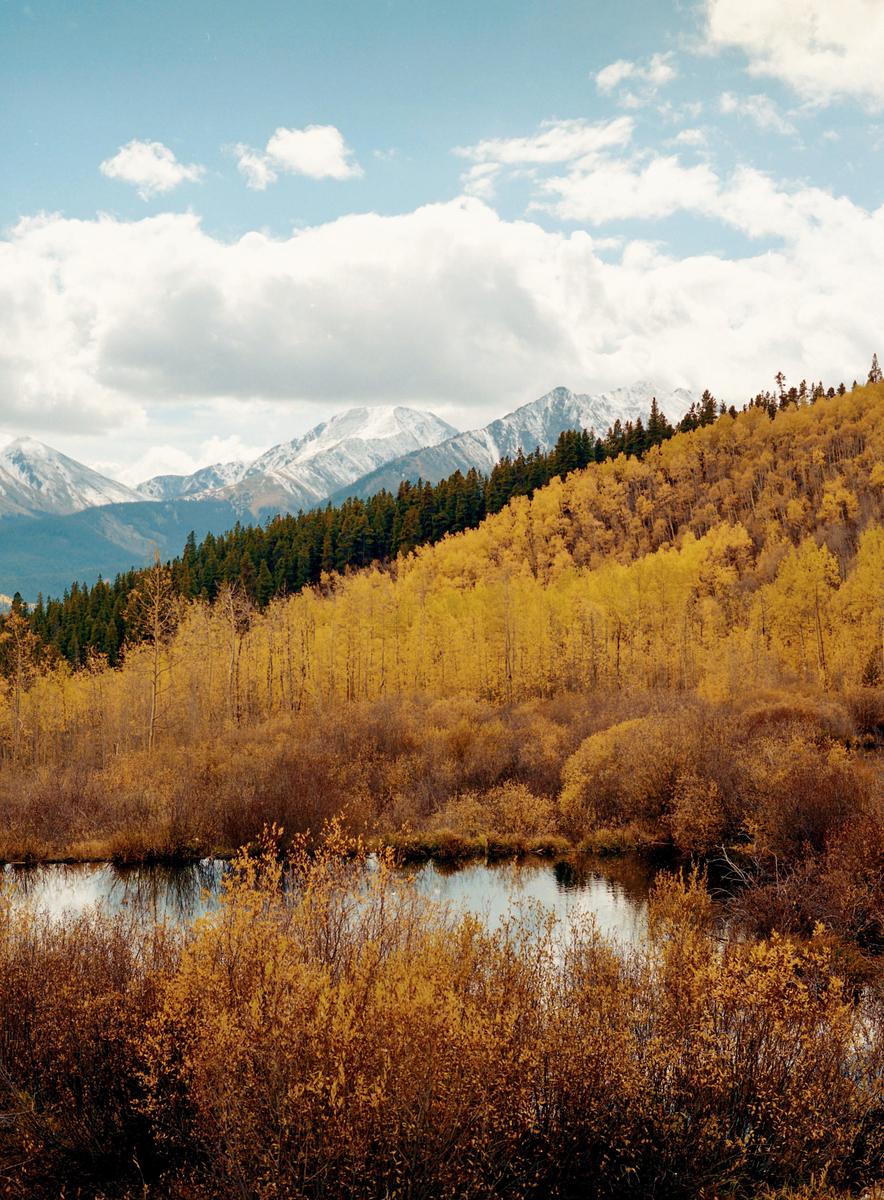 A body of water reflects the sky and is surrounded by autumn foliage with a mountain range in the background.