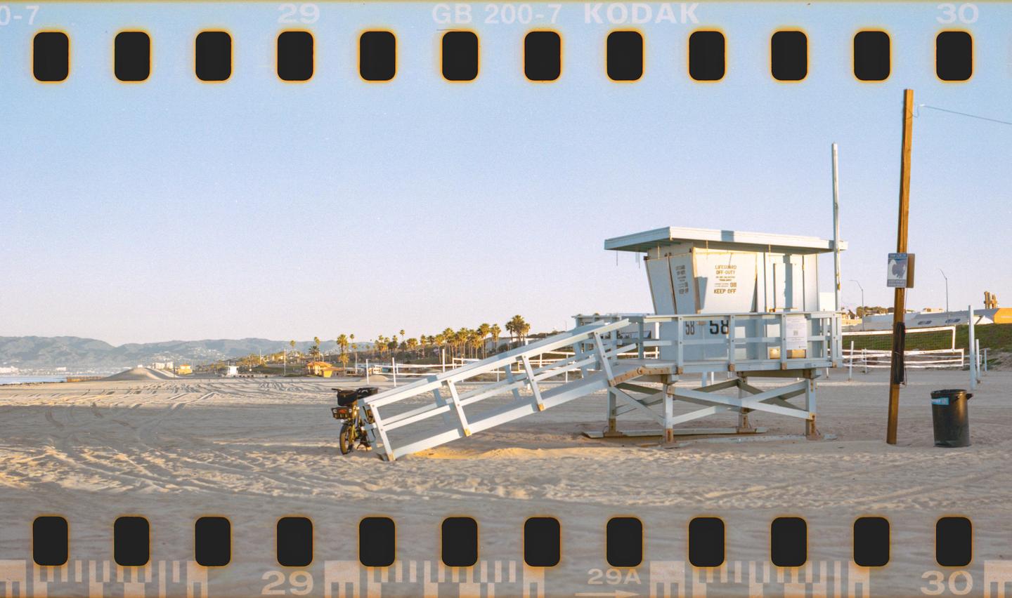 A lifeguard tower stands on a sandy beach with palm trees and mountains in the distance.