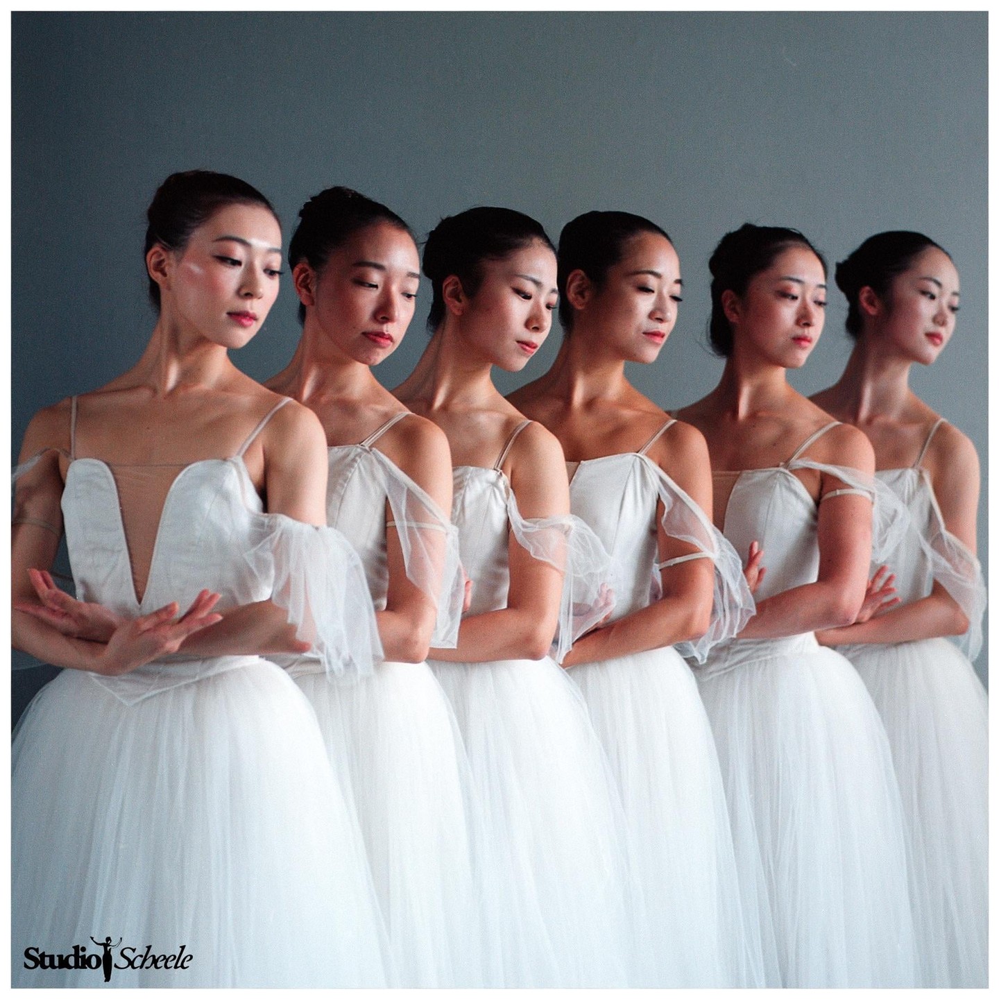 Five ballerinas in white tutus stand in a line with their arms crossed.