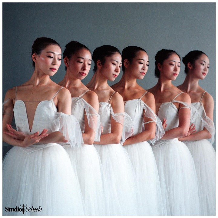 Five ballerinas in white tutus stand in a line with their arms crossed.