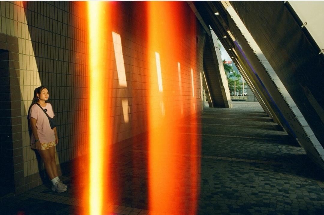 A woman stands against a tiled wall with a bright orange light leak across the frame.