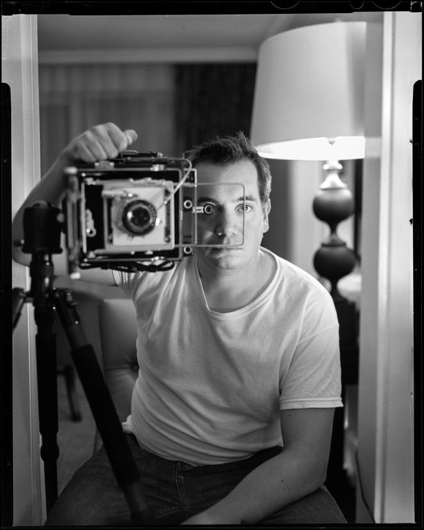 A man holds a large format camera in front of his face, taking a self-portrait in a mirror.
