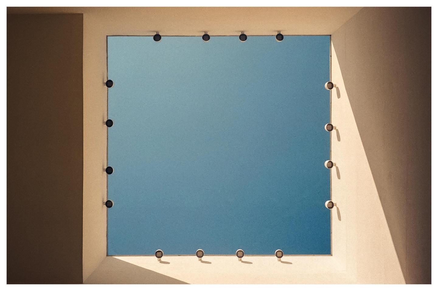A square opening in a building frames a clear blue sky, with small lights lining the edges of the opening.