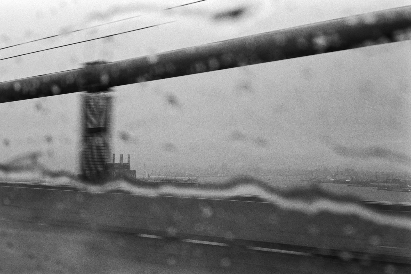 A rainy view from a moving vehicle shows a bridge railing, a distant industrial building, and a body of water with a city skyline on the horizon.