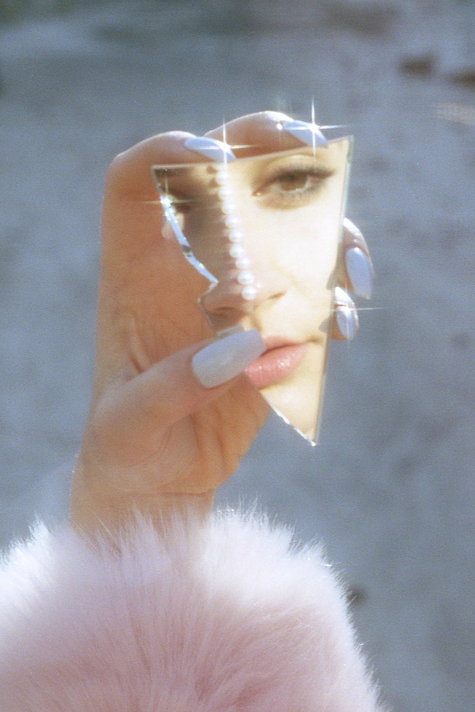 A woman's face is reflected in a broken mirror held by a hand with long nails.