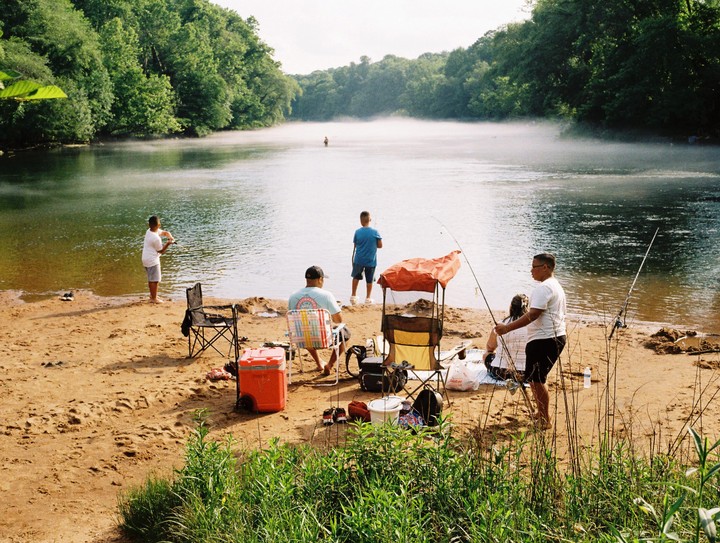 Several people are fishing on a sandy riverbank with mist rising from the water.